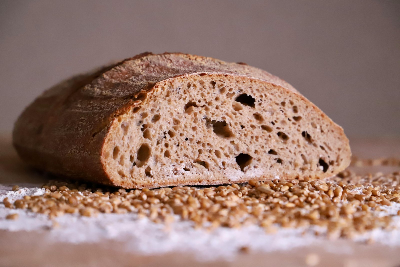 Close-up of a rustic artisan bread loaf slice surrounded by scattered wheat grains on a flour-dusted surface.